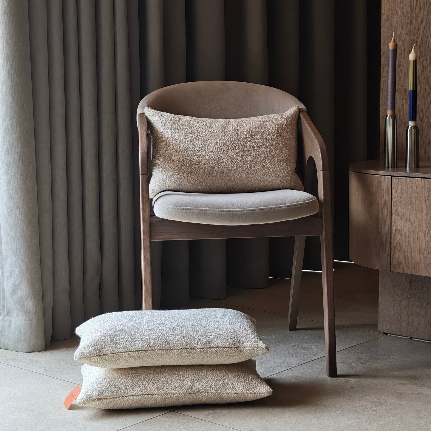 Lifestyle image of off white boucle cushions stacked on tile floors and on the chair against the armrest two stainless steel candlesticks on the rounded tv cabinet with curtains background  