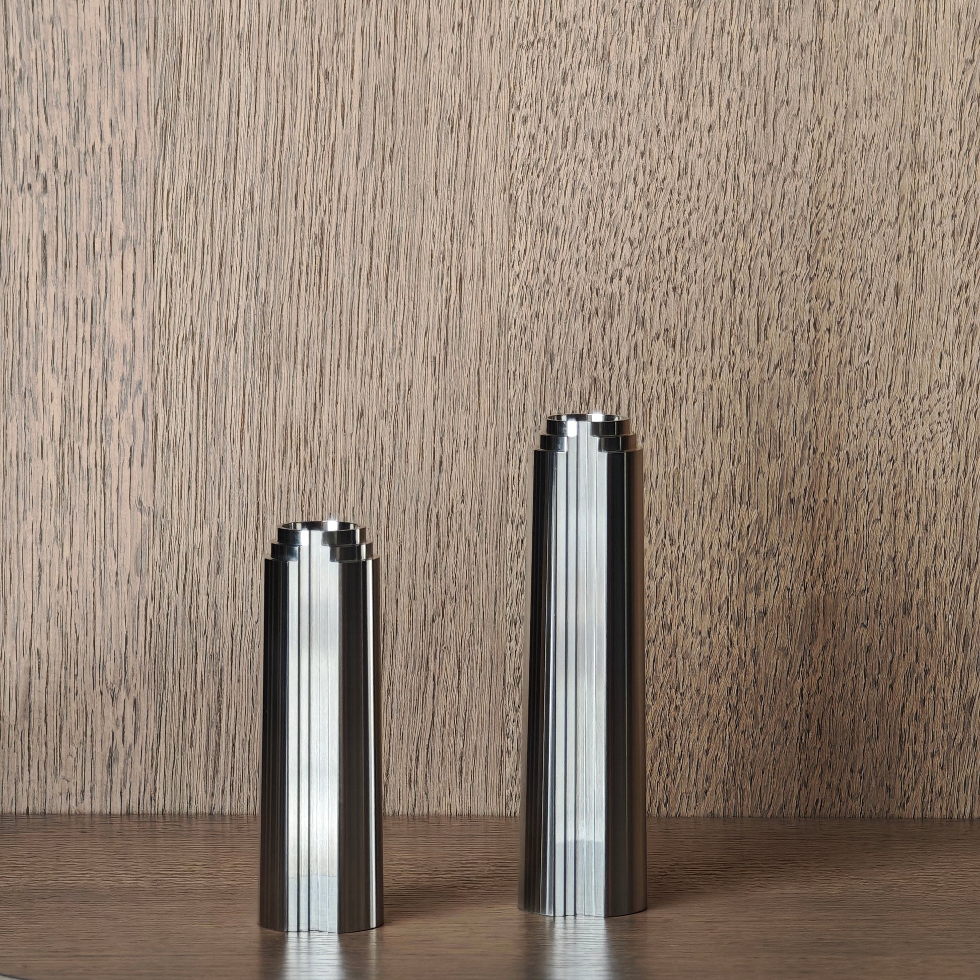 Two metallic cylindrical candle sticks on a wooden surface with a wooden wall background