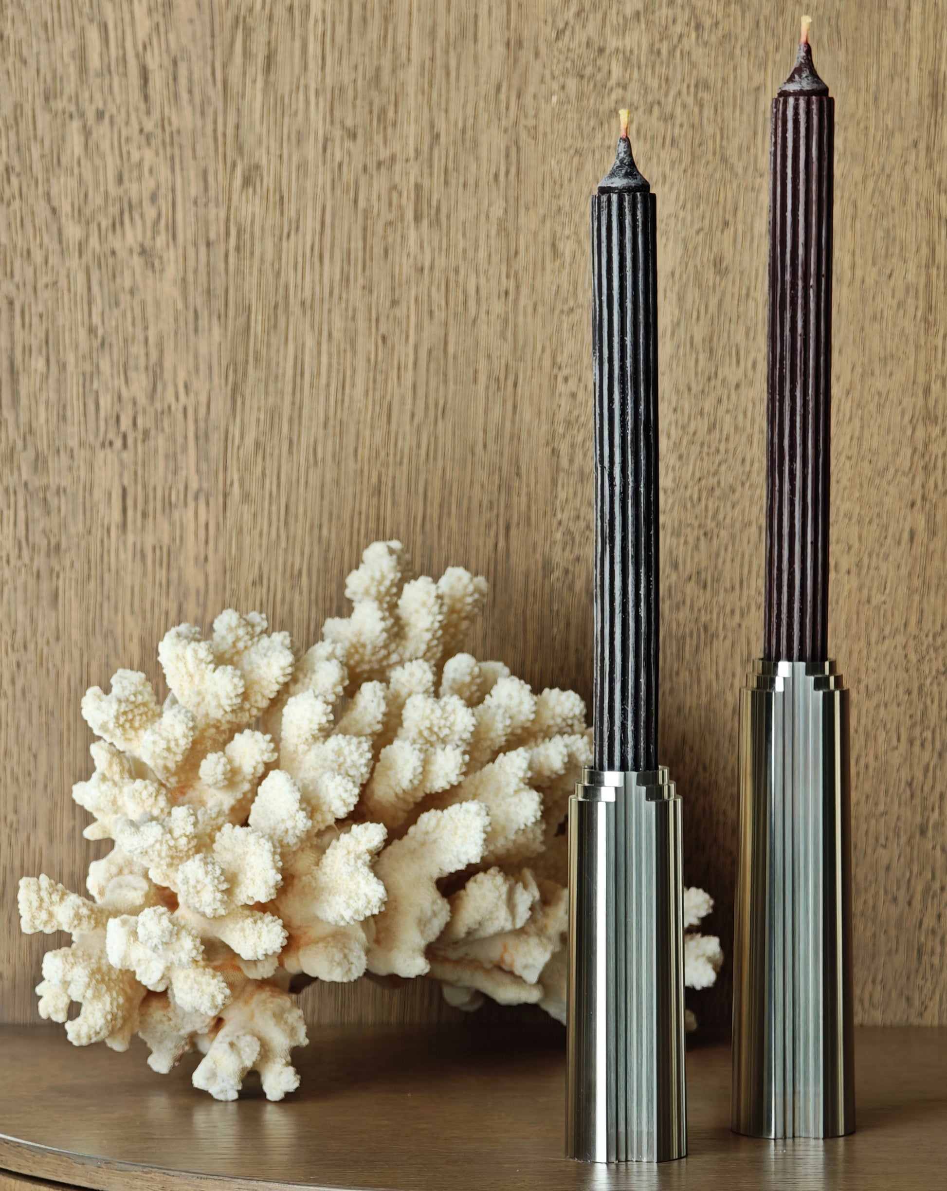 Decorative coral piece and two tall cylindrical metal candle holders on a wooden surface with a wooden wall background.