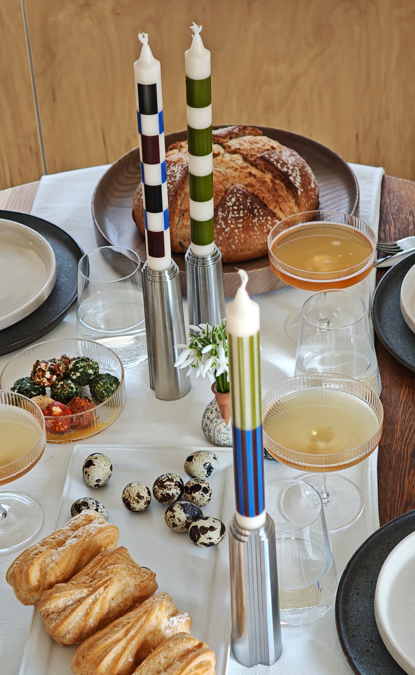 Dining table with pastries, candlesticks with colourful candles, and glasses on a white tablecloth with plywood background.