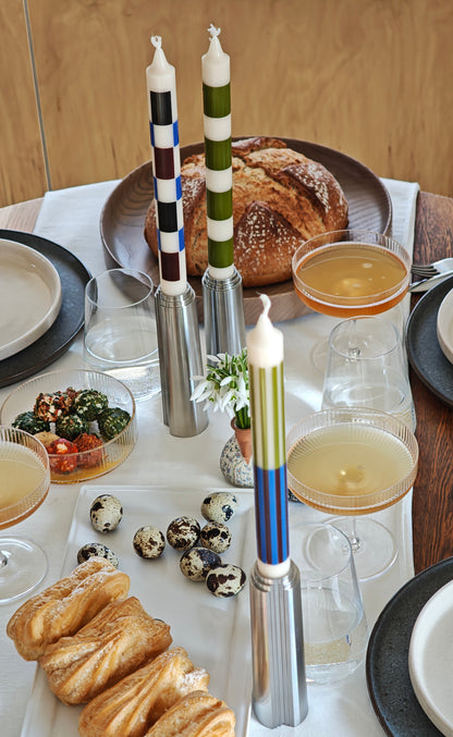 Dining table with pastries, candlesticks with colourful candles, and glasses on a white tablecloth with plywood background.
