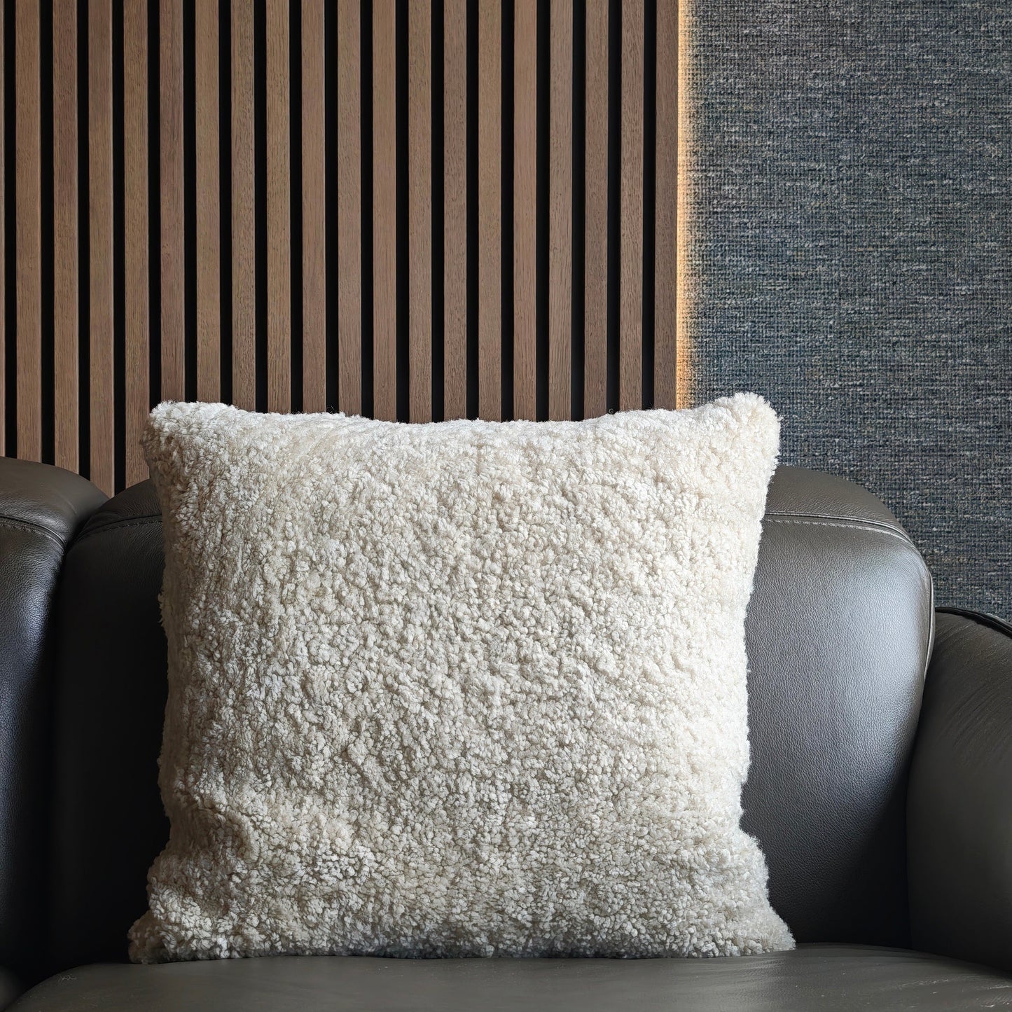 Textured white pillow on a dark grey sofa against a striped wooden slats wall and textured dark Romo wallpaper.