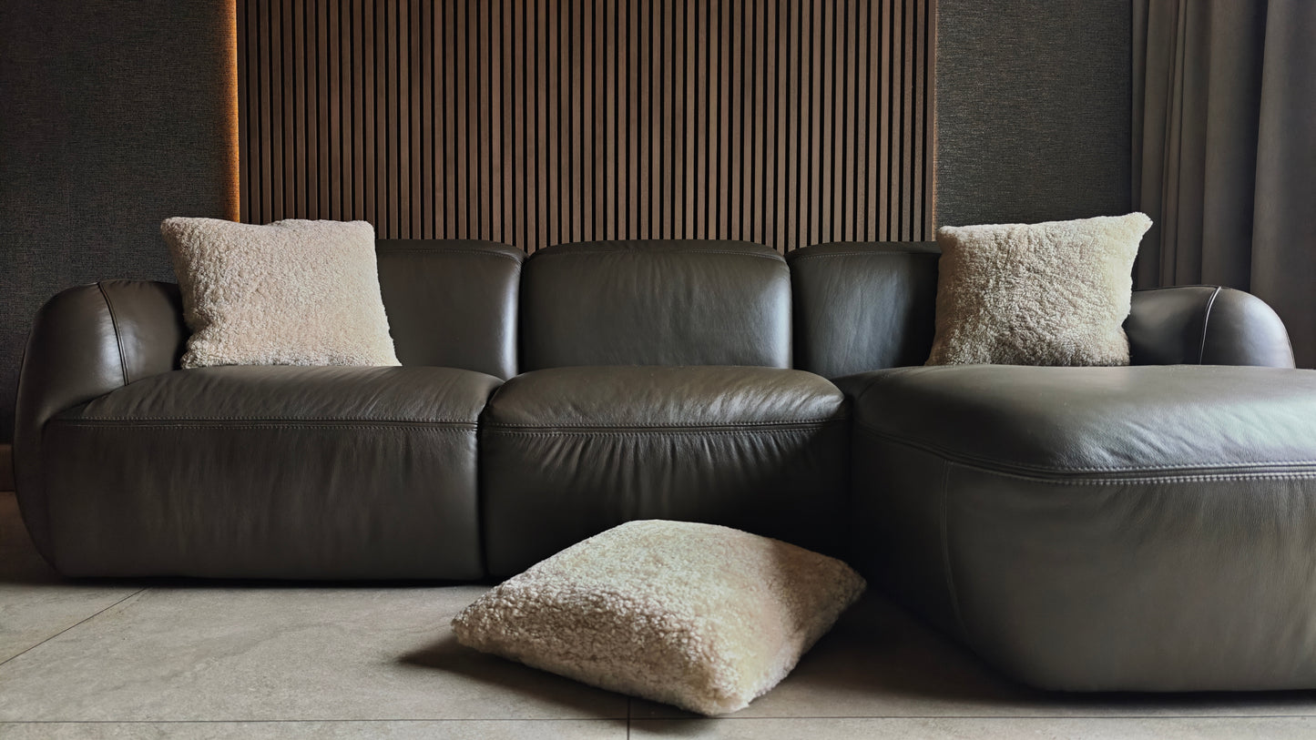 Textured off white sheepskin cushion on dark leather sofa against wooden slats paneling with wallpaper and curtains and tiles flooring