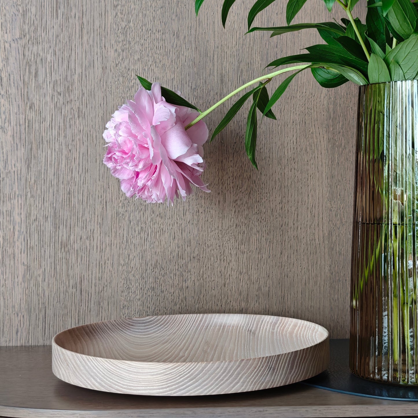 Decorative setting with a wooden tray, pink flower, and vase against a neutral wooden wall.
