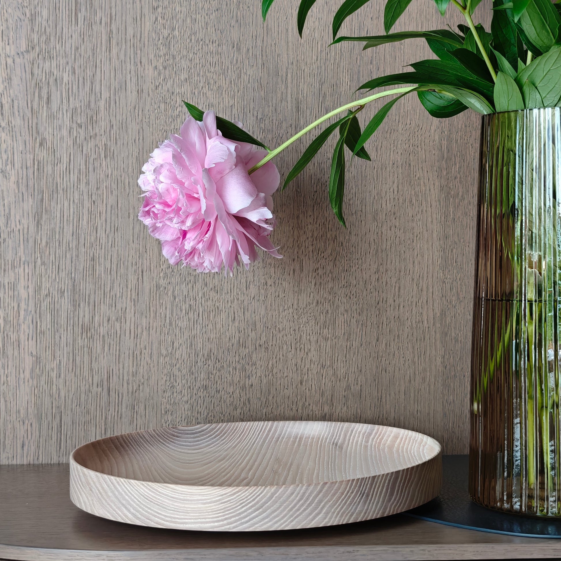 Decorative setting with a wooden tray, pink flower, and vase against a neutral wooden wall.